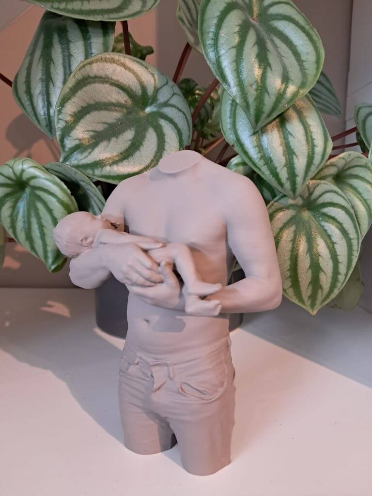 3D-beeldje van een vader die zijn liggende baby in beide armen vasthoudt, gefotografeerd voor groene kamerplanten.